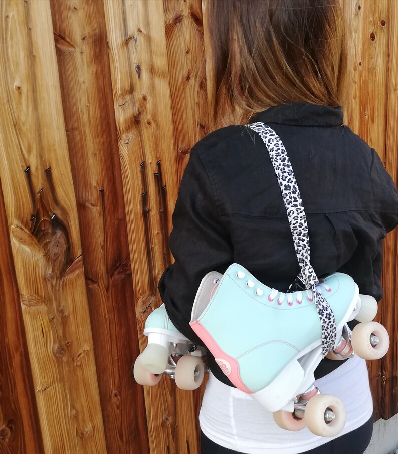May include: A person wearing a black shirt and carrying a pair of light blue roller skates with white wheels and pink accents. The skates are attached to a leopard print strap.