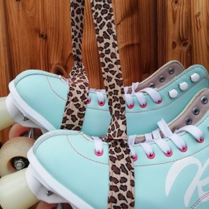 Könnte beinhalten: Ein Paar türkisfarbene Rollschuhe mit weißen Schnürsenkeln und einer weißen Basis. Die Schuhe sind mit einem braunen und schwarzen Leopardenmusterband zusammengebunden.
