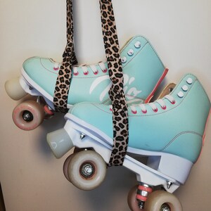 May include: A pair of teal roller skates with pink laces and white wheels. The skates are hanging from a leopard print strap.
