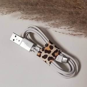 Könnte beinhalten: Weißes Ladekabel mit einem Kabelorganisator aus Leopardenmusterstoff. Das Kabel ist aufgerollt und der Stecker ist sichtbar.