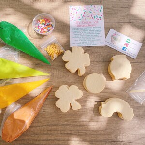 May include: DIY cookie kit with four cookie cutters, four bags of icing, sprinkles, and instructions. The cookie cutters are shaped like a shamrock, a pot of gold, a rainbow, and a circle. The icing is in green, yellow, orange, and brown.
