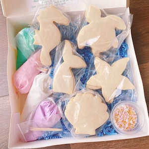 May include: A cookie decorating kit with a sea life theme. The kit includes six sugar cookies in the shapes of a seahorse, a mermaid tail, a turtle, a starfish, a seashell, and a clam. The kit also includes three bags of frosting in green, pink, and purple, a bag of sprinkles, and two wooden sticks.