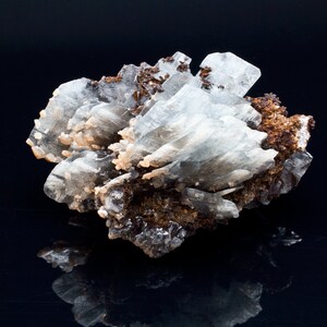 May include: A cluster of light blue crystal formations with a layered appearance, accented by patches of brown and tan minerals. The crystals are set against a dark background, creating a contrast that highlights their intricate structure.