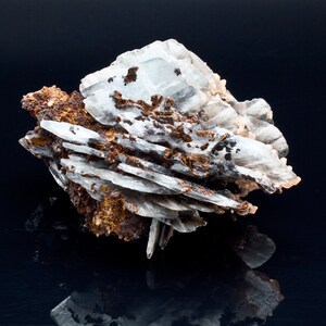 May include: A close-up of a mineral specimen featuring layered, translucent white crystals with brown and black inclusions. The mineral has a rough, textured appearance and sits against a dark, reflective surface.