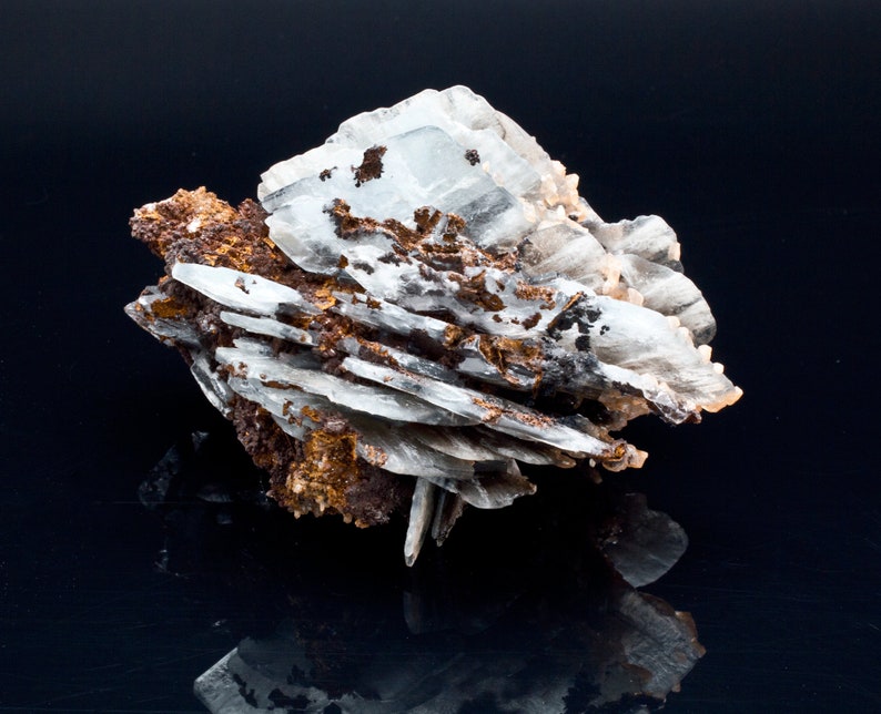 May include: A close-up of a mineral specimen featuring layered, translucent white crystals with brown and black inclusions. The mineral has a rough, textured appearance and sits against a dark, reflective surface.