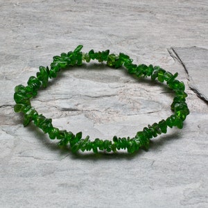 May include: A green gemstone chip bracelet made of small, irregular pieces of green stone. The bracelet is laid out on a gray, textured surface.