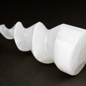 Puede incluir: Cristal de selenita blanco tallado en forma de espiral. El cristal es liso y pulido, con una apariencia ligeramente translúcida.