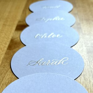 Op de afbeelding: Vijf ronde, witte, papieren plaatskaartjes met zilveren kalligrafie. De namen "Sarah", "Chloe", "Sophie", "Jill" en "Louise" staan op de kaartjes.