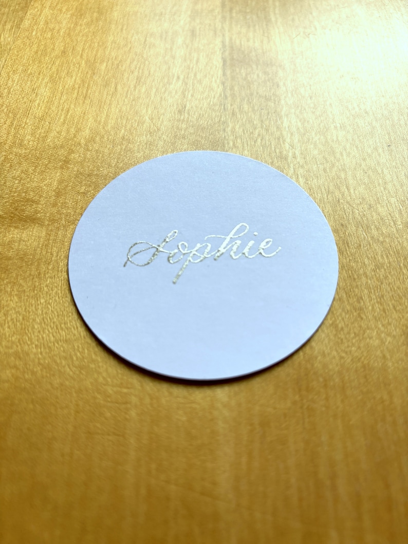 Op de afbeelding: Een ronde witte papieren plaatskaart met de naam "Sophie" geschreven in zilveren folie letters.