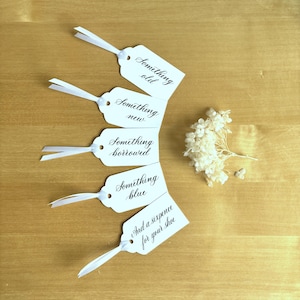 May include: Five white gift tags with black calligraphy. The tags read "Something old", "Something new", "Something borrowed", "Something blue", and "And a sixpence for your shoe".