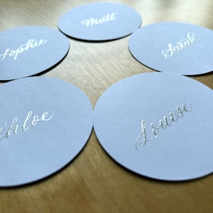 Op de afbeelding: Vijf ronde, lichtblauwe plaatskaarten met zilveren kalligrafie. De namen op de kaarten zijn Sophie, Matt, Sarah, Chloe en Louise.