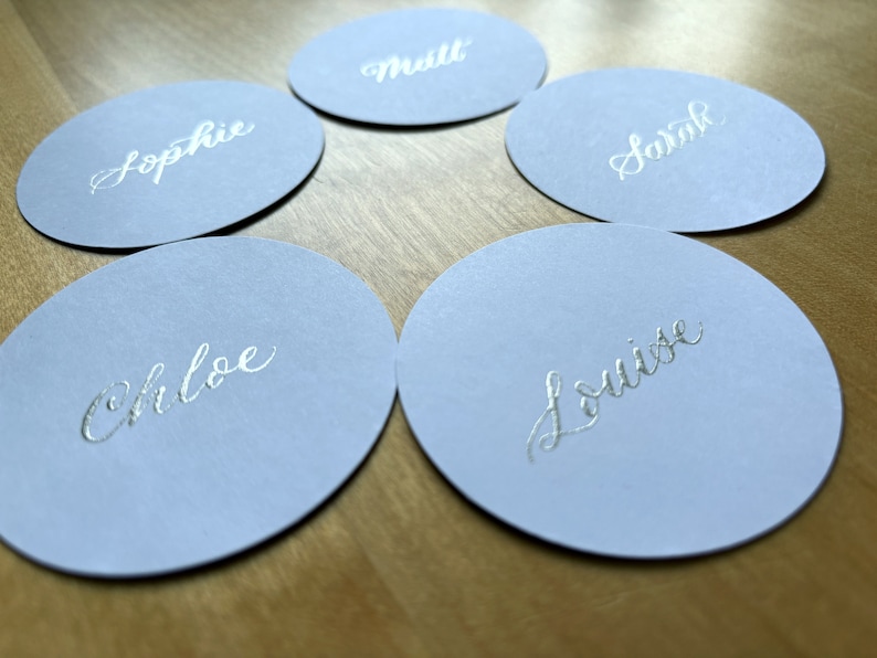 Op de afbeelding: Vijf ronde, lichtblauwe plaatskaarten met zilveren kalligrafie. De namen op de kaarten zijn Sophie, Matt, Sarah, Chloe en Louise.