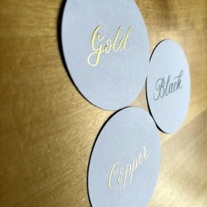 Op de afbeelding: Drie ronde, grijze papieren onderzetters met gouden letters. De onderzetters zeggen "Gold", "Black" en "Copper".