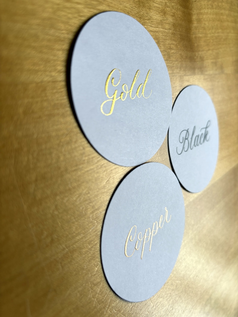 Op de afbeelding: Drie ronde, grijze papieren onderzetters met gouden letters. De onderzetters zeggen "Gold", "Black" en "Copper".