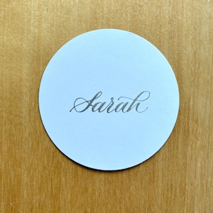 Op de afbeelding: Een ronde witte plaatskaart met de naam "Sarah" geschreven in zwarte kalligrafie.