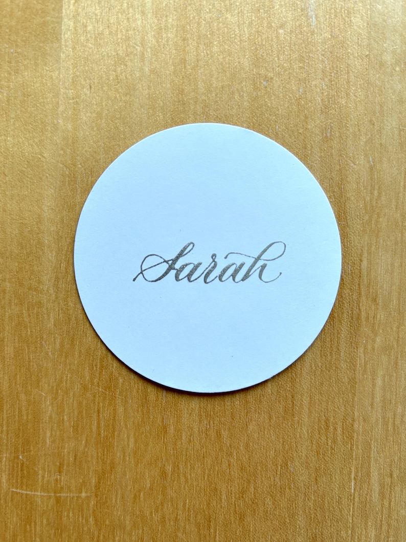 Op de afbeelding: Een ronde witte plaatskaart met de naam "Sarah" geschreven in zwarte kalligrafie.