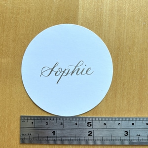 Op de afbeelding: Een ronde witte plaatskaart met de naam "Sophie" geschreven in zilveren kalligrafie.
