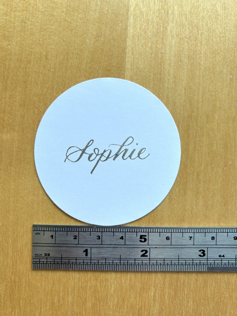 Op de afbeelding: Een ronde witte plaatskaart met de naam "Sophie" geschreven in zilveren kalligrafie.