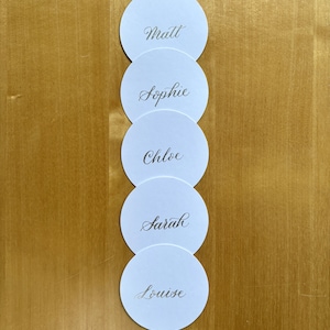 Op de afbeelding: Vijf ronde witte plaatskaarten met namen in kalligrafie. De namen zijn Matt, Sophie, Chloe, Sarah en Louise.