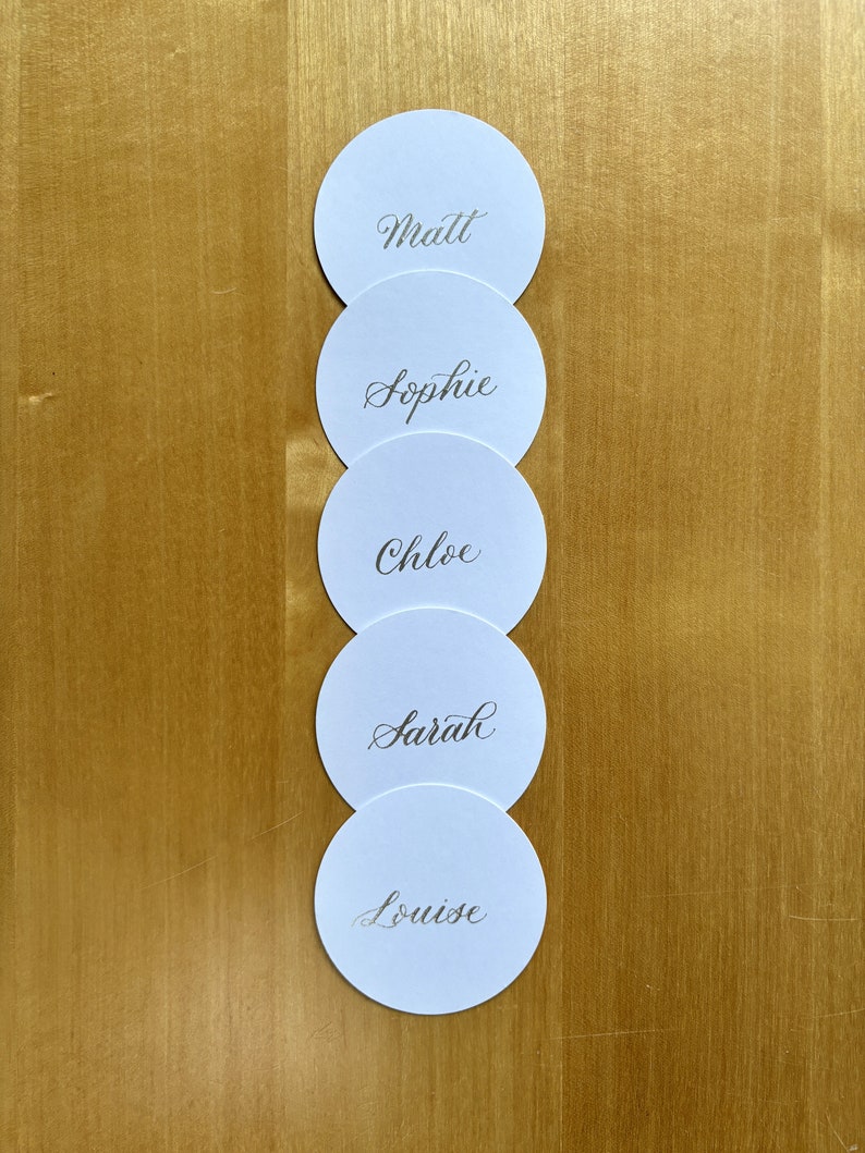 Op de afbeelding: Vijf ronde witte plaatskaarten met namen in kalligrafie. De namen zijn Matt, Sophie, Chloe, Sarah en Louise.