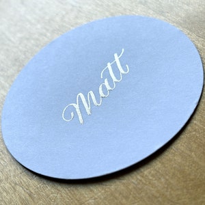 Op de afbeelding: Een ronde, lichtblauwe plaatskaart met de naam "Matt" in zilveren letters.