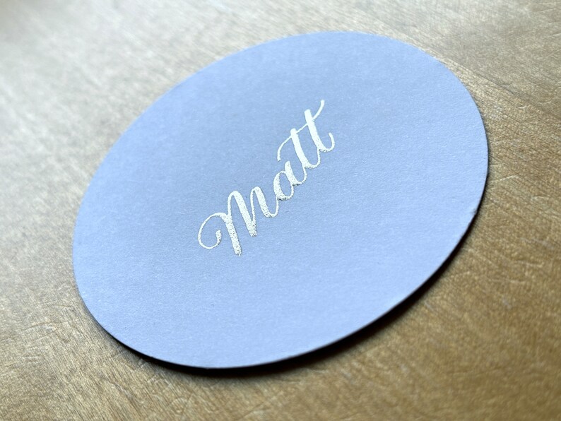 Op de afbeelding: Een ronde, lichtblauwe plaatskaart met de naam "Matt" in zilveren letters.