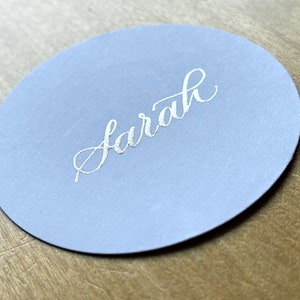 Op de afbeelding: Een ronde, lichtblauwe plaatskaart met de naam "Sarah" geschreven in zilveren kalligrafie.