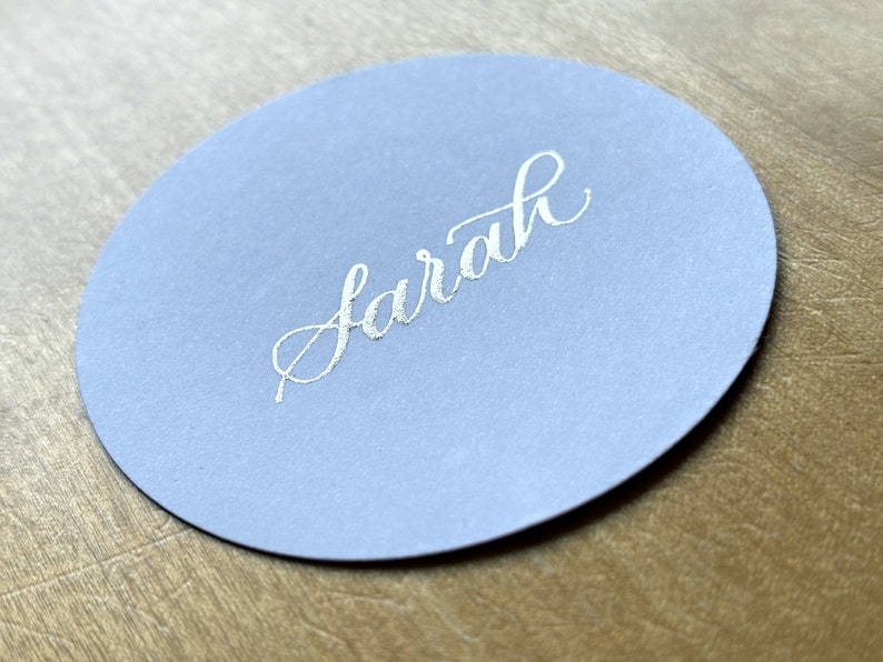 Op de afbeelding: Een ronde, lichtblauwe plaatskaart met de naam "Sarah" geschreven in zilveren kalligrafie.