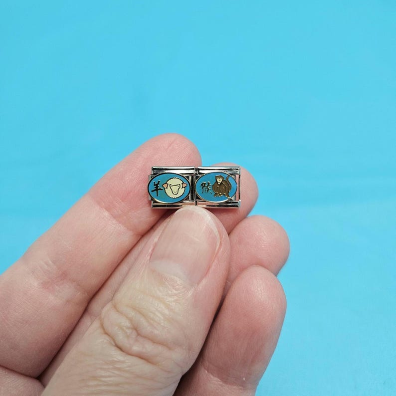May include: Two silver charm bracelets with blue enamel and Chinese characters. The left charm features a white sheep with the character "羊" for "sheep" and the right charm features a brown monkey with the character "猴" for "monkey".