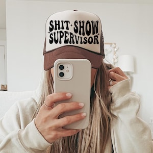 May include: A brown and white trucker hat with the text "SHIT SHOW SUPERVISOR" in black script. The hat is worn by a person holding a phone, with long blonde hair and a beige sweatshirt.
