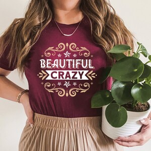Pu&ograve; includere: Una maglietta color granata con una grafica dorata e bianca che dice "Beautiful Crazy" con un bordo decorativo di stelle e volute.