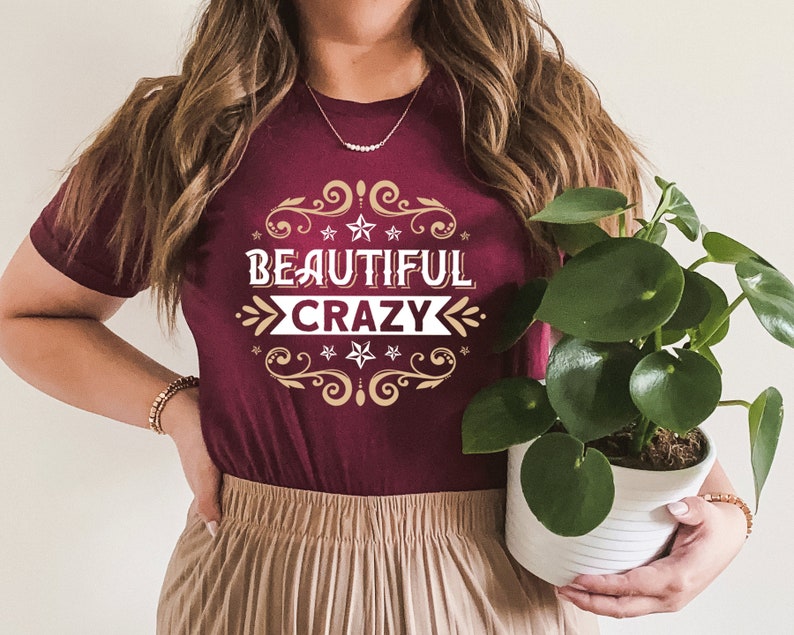 Pu&ograve; includere: Una maglietta color granata con una grafica dorata e bianca che dice "Beautiful Crazy" con un bordo decorativo di stelle e volute.