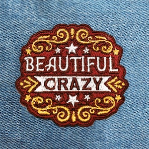 May include: Embroidered patch with the words "BEAUTIFUL" and "CRAZY" in white on a maroon background. The patch has gold decorative swirls and stars. The patch is on a blue denim background.