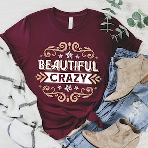 Pu&ograve; includere: Maglietta color borgogna con un design oro e bianco che dice "Beautiful Crazy".