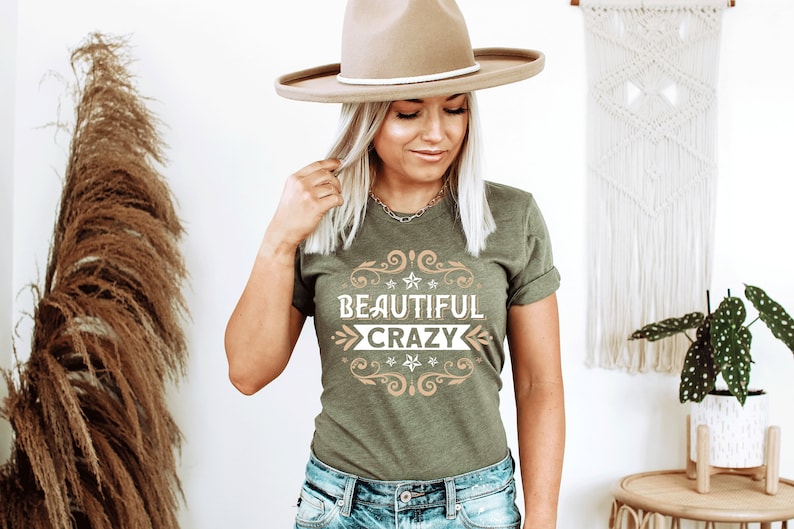 Pu&ograve; includere: Una donna indossa una maglietta verde con il testo "Beautiful Crazy" in un font decorativo. Indossa un cappello marrone e jeans blu.