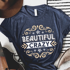 Pu&ograve; includere: Una t-shirt blu navy con una grafica bianca che dice "Beautiful Crazy" in un font vintage con accenti dorati e disegni floreali.