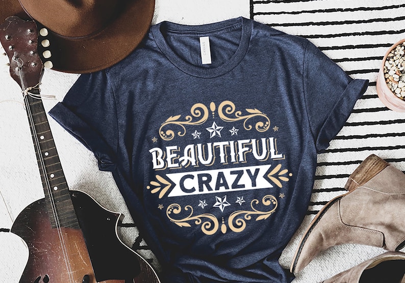 Pu&ograve; includere: Una t-shirt blu navy con una grafica bianca che dice "Beautiful Crazy" in un font vintage con accenti dorati e disegni floreali.