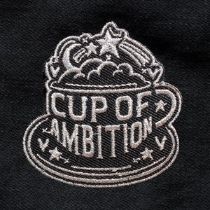 May include: A black fabric patch featuring white embroidery of a coffee cup with the text CUP OF AMBITION. The design includes a crescent moon, stars, and clouds rising from the cup, all rendered in white thread against a solid black background.