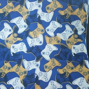May include: A blue fabric with a repeating pattern of white and black video game controllers. The controllers are outlined in gold.