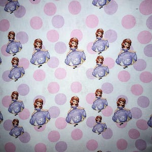 May include: A patterned fabric featuring a cartoon princess in a purple dress, with brown hair and a tiara. The background is white with pink and purple polka dots. Suitable for children's crafts or clothing. The fabric is likely cotton.
