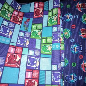 May include: Fabric featuring a colorful pattern with characters from the PJ Masks animated series. The design includes images of Catboy, Owlette, and Gekko in various poses, set against a blue, purple, and green background.