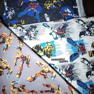 Puede incluir: Tres piezas de tela con personajes de Transformers. Una pieza es negra con un patrón hexagonal, una es blanca con detalles de puntos azules y el logotipo de Transformers, y una es gris. Todas las telas muestran varias figuras de robots.