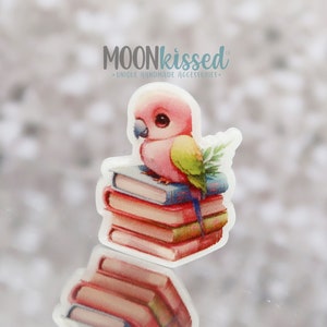 Pode incluir: Um autocolante com um papagaio rosa empoleirado numa pilha de livros. O papagaio tem uma asa verde e amarela. Os livros são vermelhos e azuis. O autocolante tem o texto "MOONKISSED" e "UNIQUE HANDMADE ACCESSORIES".