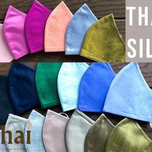 May include: A collection of 12 face masks made from Thai silk in various colors, including pink, purple, peach, blue, green, and brown. The masks are arranged in two rows on a wooden surface. The text "THAI SILK" is displayed in a white box on a gray background.