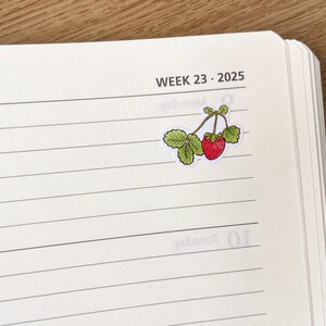 May include: A white page with black lines and the text "WEEK 23 - 2025" at the top. A sticker of a red strawberry with green leaves is on the page.