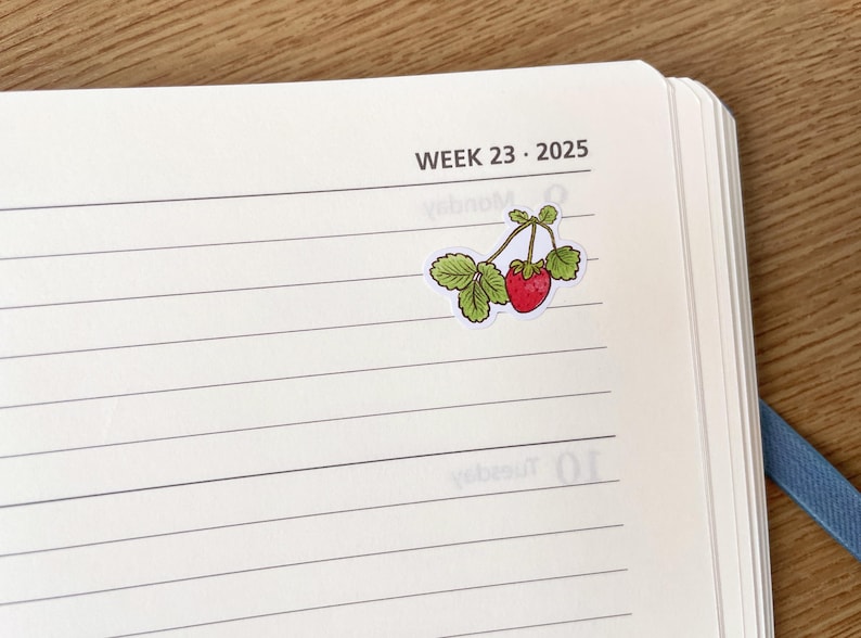 May include: A white page with black lines and the text "WEEK 23 - 2025" at the top. A sticker of a red strawberry with green leaves is on the page.
