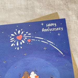 Może przedstawiać: Niebieska kartka z białym wzorem fajerwerk&oacute;w i dwoma czerwonymi sercami. Na kartce napis "Happy Anniversary" w srebrnym brokacie.