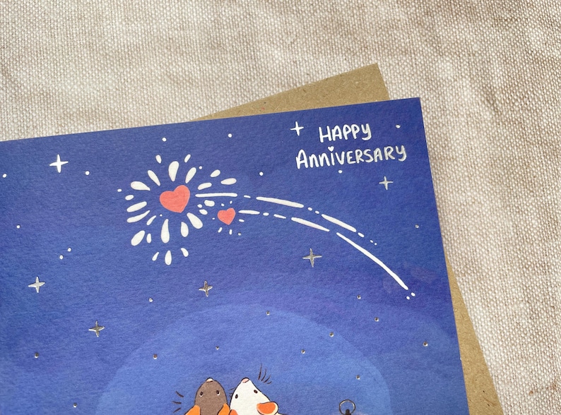 Może przedstawiać: Niebieska kartka z białym wzorem fajerwerk&oacute;w i dwoma czerwonymi sercami. Na kartce napis "Happy Anniversary" w srebrnym brokacie.