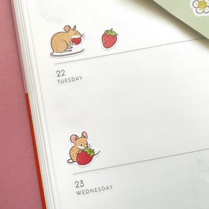 May include: A planner with two stickers of a mouse eating a strawberry. The planner has the numbers 22 and 23 with the days Tuesday and Wednesday written below them.
