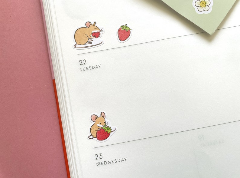 May include: A planner with two stickers of a mouse eating a strawberry. The planner has the numbers 22 and 23 with the days Tuesday and Wednesday written below them.